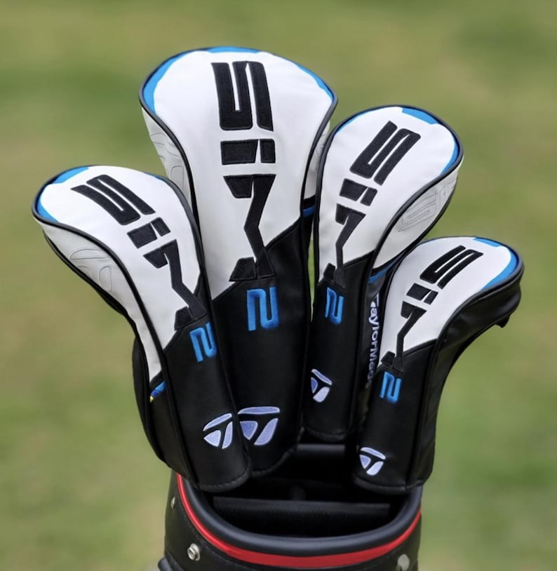 May include: A set of four golf club head covers in a golf bag. The covers are white with black and blue accents, featuring the number "2" and the letters "SIF" in black. The TaylorMade logo is visible.