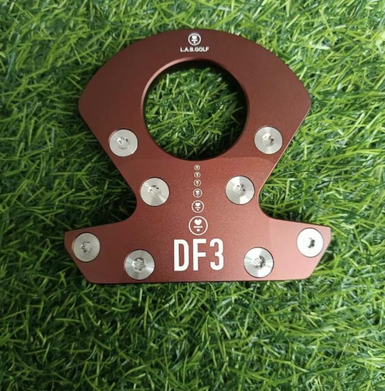 Op de afbeelding: Een roodbruin golfclubaccessoire met een centraal gat en de tekst "DF3" in het wit. Het accessoire heeft meerdere zilveren schroeven en het logo "L.A.B. GOLF" bovenaan. Het item staat op een groene grasachtergrond.