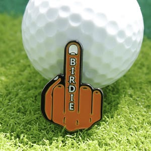 Könnte beinhalten: Ein Golfball mit einem orangefarbenen und schwarzen Birdie-Finger-Ballmarker. Der Marker hat die Form einer Hand mit ausgestrecktem Mittelfinger, und das Wort "Birdie" ist vertikal geschrieben.
