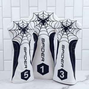 Op de afbeelding: Een set van drie witte golfclubhoofdbeschermers met een zwart spinnenwebontwerp. Het woord "SPIDER" staat verticaal op elke beschermer. De nummers 1, 3 en 5 worden weergegeven.