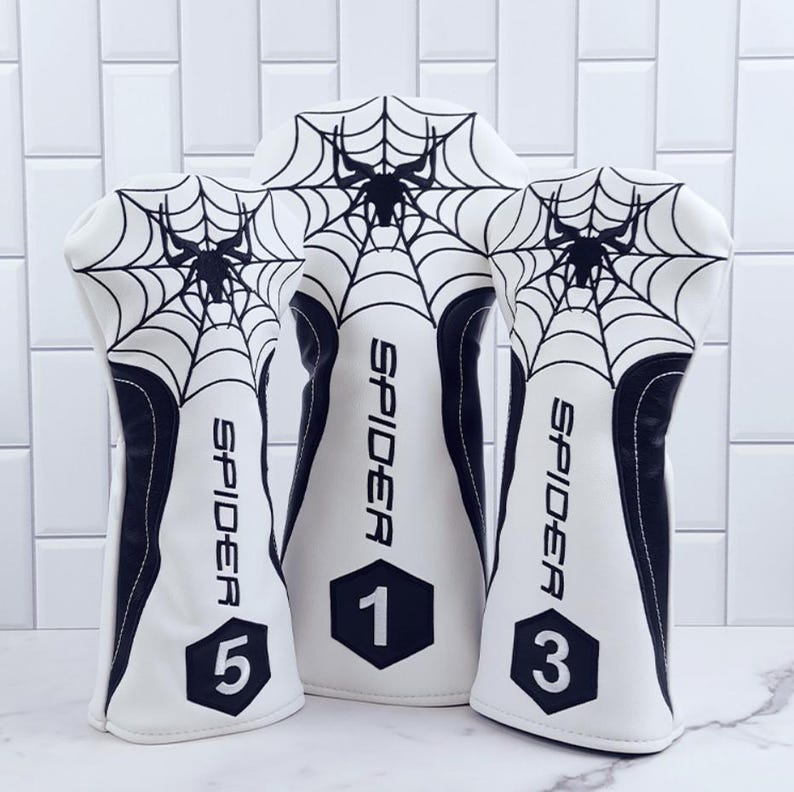 Op de afbeelding: Een set van drie witte golfclubhoofdbeschermers met een zwart spinnenwebontwerp. Het woord "SPIDER" staat verticaal op elke beschermer. De nummers 1, 3 en 5 worden weergegeven.