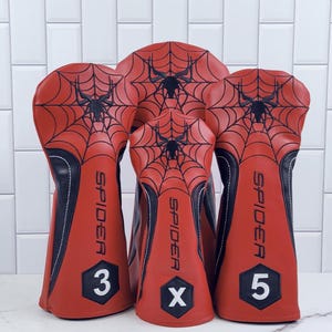 Op de afbeelding: Set van vier rode en zwarte golfclubhoofdbedekkingen. Elke hoes heeft een zwart spinnenwebontwerp op het rode oppervlak. Het woord "SPIDER" staat verticaal op elke hoes gedrukt. De cijfers 3 en 5 en de letter X staan op de hoezen.