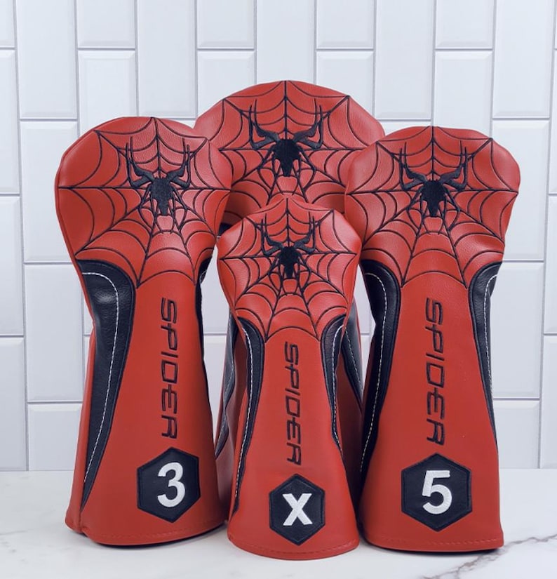 Op de afbeelding: Set van vier rode en zwarte golfclubhoofdbedekkingen. Elke hoes heeft een zwart spinnenwebontwerp op het rode oppervlak. Het woord "SPIDER" staat verticaal op elke hoes gedrukt. De cijfers 3 en 5 en de letter X staan op de hoezen.