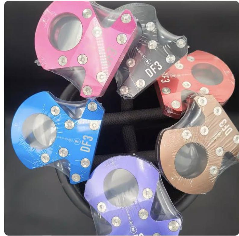 Op de afbeelding: Een verzameling kleurrijke metalen hardware-onderdelen, waaronder roze, zwart, rood, brons, blauw en paars. Elk stuk heeft een cirkelvormige opening en is vastgezet met zilveren schroeven. De tekst "DF3" is zichtbaar op sommige stukken.