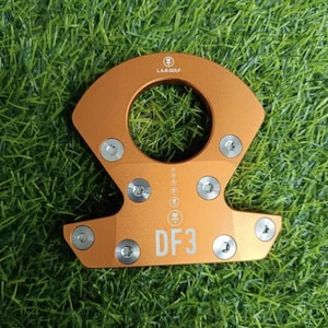 Op de afbeelding: Oranje metalen golfclub putterkop met de tekst "DF3" en "LAB GOLF". De putterkop heeft een cirkelvormige opening en is bevestigd met zilveren schroeven. De putterkop staat op een groene grasachtergrond.
