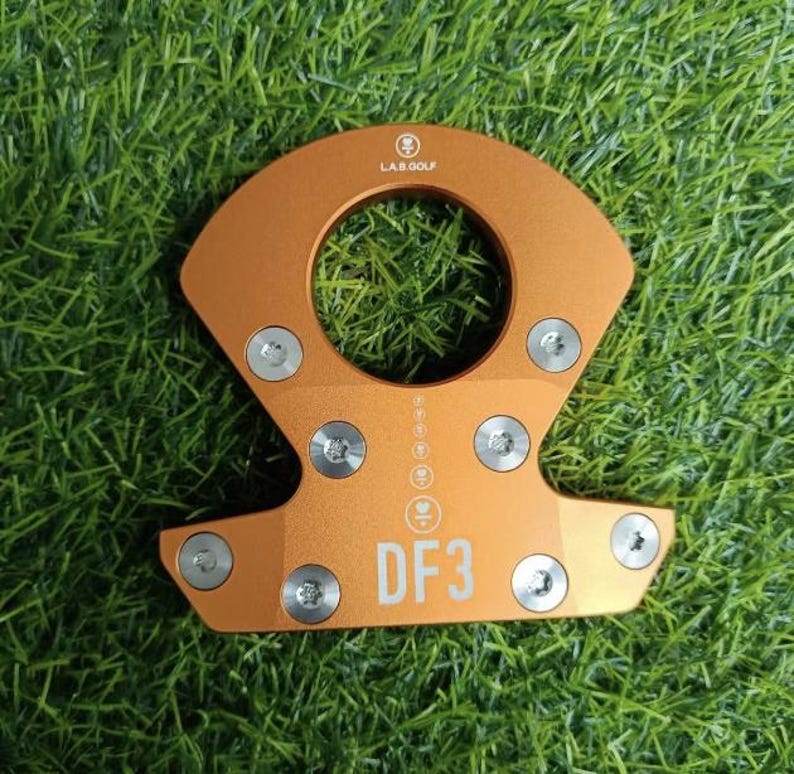 Op de afbeelding: Oranje metalen golfclub putterkop met de tekst "DF3" en "LAB GOLF". De putterkop heeft een cirkelvormige opening en is bevestigd met zilveren schroeven. De putterkop staat op een groene grasachtergrond.