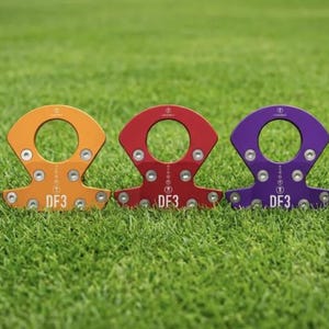 Op de afbeelding: Vijf kleurrijke metalen trainingshulpmiddelen zijn gerangschikt op een groene grasachtige ondergrond. Elk hulpmiddel heeft een cirkelvormige opening en een basis met meerdere schroefgaten. De kleuren zijn blauw, oranje, rood, paars en bruin. Elk hulpmiddel heeft de tekst "DF3".