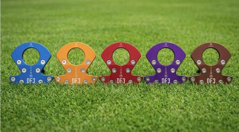 Op de afbeelding: Vijf kleurrijke metalen trainingshulpmiddelen zijn gerangschikt op een groene grasachtige ondergrond. Elk hulpmiddel heeft een cirkelvormige opening en een basis met meerdere schroefgaten. De kleuren zijn blauw, oranje, rood, paars en bruin. Elk hulpmiddel heeft de tekst "DF3".