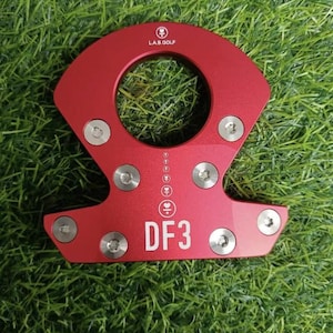 Op de afbeelding: Rood metalen golfclubaccessoire met de tekst "DF3" en "L.A.B. GOLF". Het accessoire heeft een cirkelvormige opening en is bevestigd met meerdere zilveren schroeven. Het item ligt op een groene, grasachtige ondergrond.