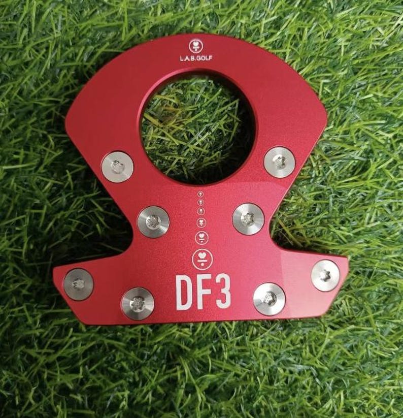 Op de afbeelding: Rood metalen golfclubaccessoire met de tekst "DF3" en "L.A.B. GOLF". Het accessoire heeft een cirkelvormige opening en is bevestigd met meerdere zilveren schroeven. Het item ligt op een groene, grasachtige ondergrond.