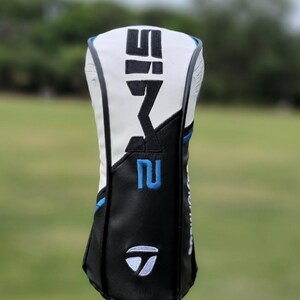 May include: A golf club head cover with a white top and black bottom. The cover has the text "SIM2" in black and the number "2" in blue. The TaylorMade logo is visible on the bottom.