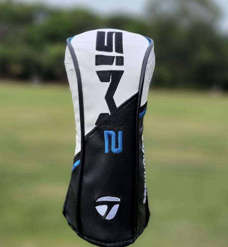 May include: A golf club head cover with a white top and black bottom. The cover has the text "SIM2" in black and the number "2" in blue. The TaylorMade logo is visible on the bottom.