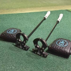 Op de afbeelding: Twee zwarte golfputters met witte grips en bijpassende zwarte headcovers. De putters hebben een cirkelvormig ontwerp en de headcovers hebben een blauw logo. De putters liggen op een groene ondergrond.