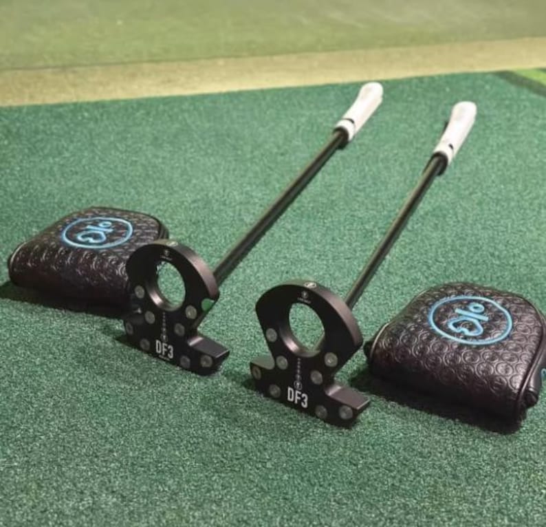 Op de afbeelding: Twee zwarte golfputters met witte grips en bijpassende zwarte headcovers. De putters hebben een cirkelvormig ontwerp en de headcovers hebben een blauw logo. De putters liggen op een groene ondergrond.