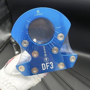 Op de afbeelding: Een blauwe golf putter met de tekst "L.A.B. GOLF" en "DF3" erop gedrukt. De putter heeft een unieke vorm met een cirkelvormige opening en is vastgezet met meerdere zilveren schroeven. Het is verpakt in doorzichtig plastic.