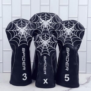 Op de afbeelding: Zwarte golfclubhoezen met een wit spinnenweb- en spinnendesign. Het woord "SPIDER" staat verticaal op elke hoes, samen met de cijfers 3, 5 en de letter X. Witte stiksels accentueren de randen.