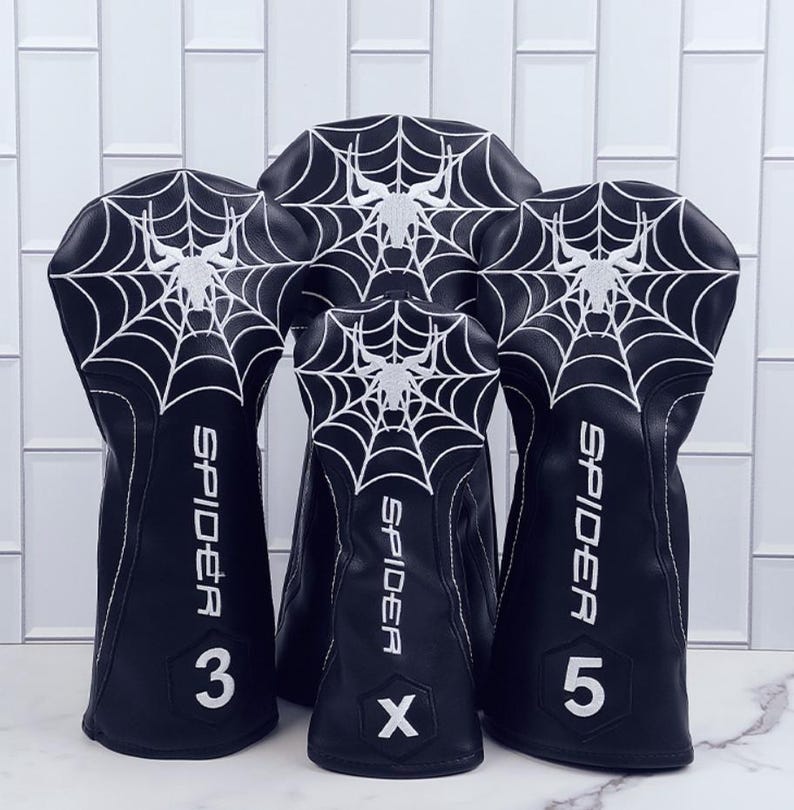 Op de afbeelding: Zwarte golfclubhoezen met een wit spinnenweb- en spinnendesign. Het woord "SPIDER" staat verticaal op elke hoes, samen met de cijfers 3, 5 en de letter X. Witte stiksels accentueren de randen.