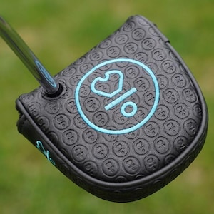 Pode incluir: Capa preta para a cabeça do putter de golfe com um logotipo circular turquesa e um padrão repetitivo. A capa está em um taco de golfe com um eixo prateado. O fundo é grama verde desfocada.