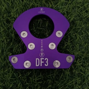 Op de afbeelding: Een paars golfclubaccessoire met de tekst "DF3" en het "LAB GOLF"-logo. Het accessoire heeft een cirkelvormige opening en is bevestigd met meerdere zilveren schroeven. Het object ligt op een groene ondergrond.