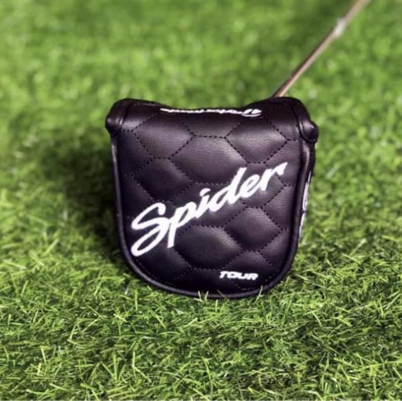 Puede incluir: Funda negra para la cabeza del palo de golf con la inscripci&oacute;n blanca "Spider" y costuras de diamante. La funda est&aacute; sobre una superficie verde y el palo de golf es parcialmente visible. La palabra "TOUR" est&aacute; impresa en blanco en la parte inferior de la funda.