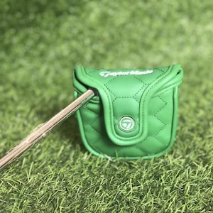 Puede incluir: Funda de putter de golf verde con un patr&oacute;n de costura de diamante. La funda tiene un cierre magn&eacute;tico y el logotipo de TaylorMade en blanco. Un palo de golf est&aacute; parcialmente insertado en la funda, sobre hierba verde.