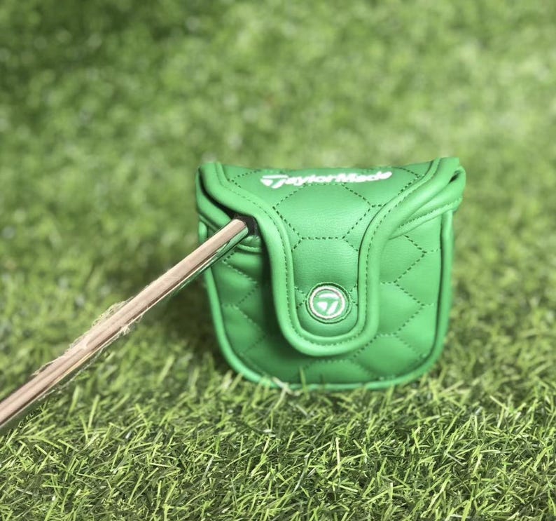Puede incluir: Funda de putter de golf verde con un patr&oacute;n de costura de diamante. La funda tiene un cierre magn&eacute;tico y el logotipo de TaylorMade en blanco. Un palo de golf est&aacute; parcialmente insertado en la funda, sobre hierba verde.