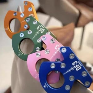 Op de afbeelding: Close-up van vier kleurrijke metalen karabijnhaken in oranje, groen, roze en blauw. Elke karabijnhaak heeft een cirkelvormige opening en een rechthoekige bovenkant met de tekst "DF3" en verschillende kleine zilveren schroeven. De karabijnhaken zijn schuin gestapeld.