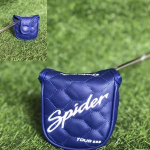 Puede incluir: Una funda de putter de golf azul acolchada con la palabra "Spider" en escritura blanca. La funda est&aacute; sobre una superficie de hierba verde. El putter est&aacute; parcialmente insertado en la funda.