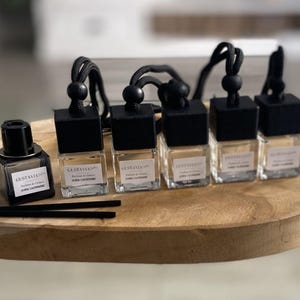 Peut inclure: Plusieurs désodorisants pour voiture avec des bouchons et des cordons noirs, présentés sur un plateau en bois. Les flacons en verre contiennent un liquide clair et sont étiquetés avec la marque "GUSTAVIA" et les détails du parfum. Deux bâtonnets diffuseurs noirs sont également présents.