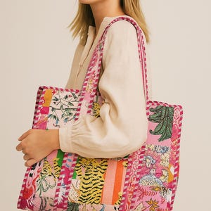 May include: A pink tote bag with a patchwork design featuring floral, tiger, and geometric patterns. The bag has a rectangular shape with a contrasting pink trim and handles. The bag is held by a person wearing a cream-colored blouse.