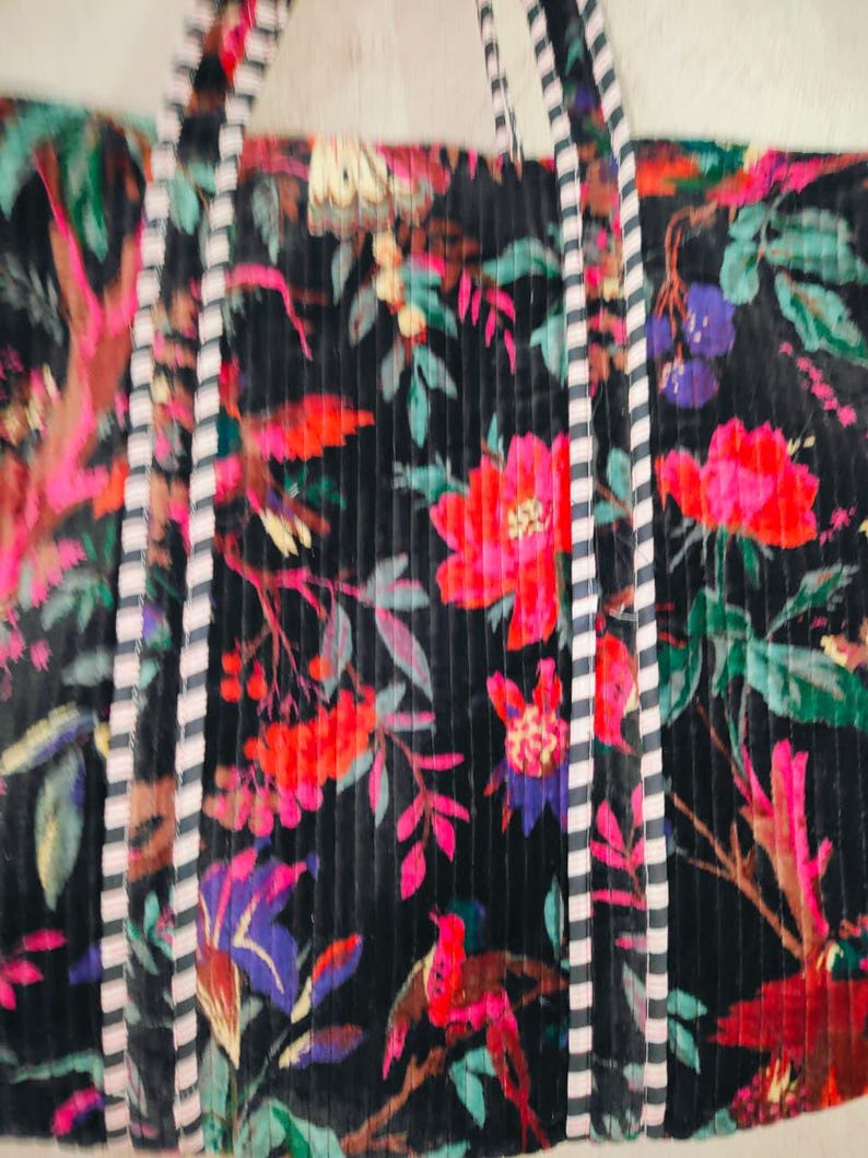 Puede incluir: Un bolso tote negro con un vibrante estampado floral con flores rojas, rosas y moradas, hojas verdes y p&aacute;jaros. El bolso tiene asas y ribetes a rayas.