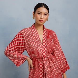 Op de afbeelding: Een rood en crèmekleurig badjas met patroon en een ceintuur. De badjas heeft verschillende patronen, waaronder stippen en geometrische vormen. Gemaakt van een zachte, soepele stof en heeft wijde mouwen.