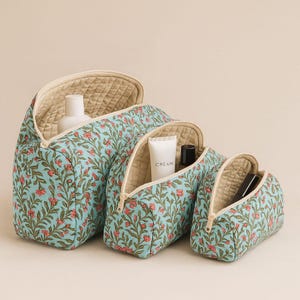 Puede incluir: Juego de tres neceseres con estampado floral de diferentes tamaños. Las bolsas tienen un fondo azul claro con un estampado floral rojo y verde. Cada bolsa tiene un forro acolchado beige y cierre de cremallera.