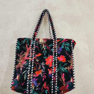 Puede incluir: Un bolso tote negro con un vibrante estampado floral en tonos rosa, naranja y verde. El bolso presenta asas y ribetes a rayas. El dise&ntilde;o incluye p&aacute;jaros y follaje, creando un accesorio colorido y llamativo.