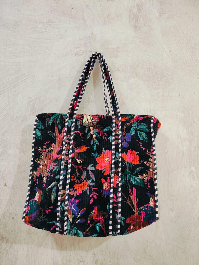 Puede incluir: Un bolso tote negro con un vibrante estampado floral en tonos rosa, naranja y verde. El bolso presenta asas y ribetes a rayas. El dise&ntilde;o incluye p&aacute;jaros y follaje, creando un accesorio colorido y llamativo.