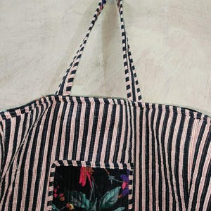 Puede incluir: Un bolso tote a rayas blancas y negras con detalles en rojo. El bolso tiene un bolsillo con estampado floral. Las asas tambi&eacute;n son a rayas. El bolso est&aacute; colgado contra una pared de color claro.