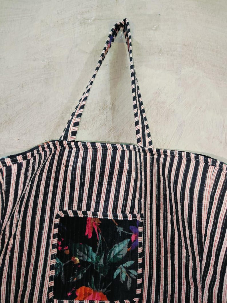 Puede incluir: Un bolso tote a rayas blancas y negras con detalles en rojo. El bolso tiene un bolsillo con estampado floral. Las asas tambi&eacute;n son a rayas. El bolso est&aacute; colgado contra una pared de color claro.