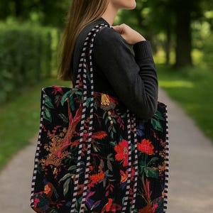 Puede incluir: Un bolso tote negro grande con un vibrante estampado floral en rojo, verde y amarillo. El bolso tiene asas a rayas y una textura acolchada. La persona lleva un su&eacute;ter gris oscuro y vaqueros azules.