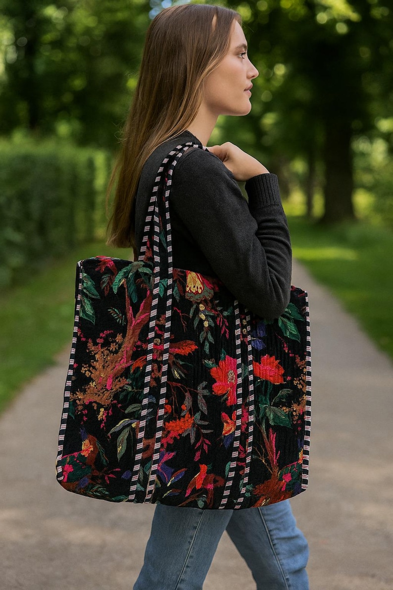 Puede incluir: Un bolso tote negro grande con un vibrante estampado floral en rojo, verde y amarillo. El bolso tiene asas a rayas y una textura acolchada. La persona lleva un su&eacute;ter gris oscuro y vaqueros azules.