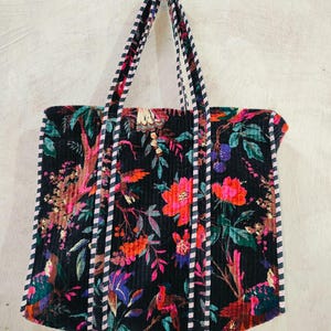 Puede incluir: Bolso tote negro con un vibrante estampado floral en tonos rojos, rosas y morados. El bolso tiene asas y ribetes a rayas. El dise&ntilde;o incluye p&aacute;jaros y follaje.