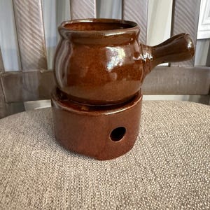 May include: A brown ceramic fondue pot with a single handle resting on a matching cylindrical base. The base features a circular opening on the side for a heat source. The entire piece has a glossy, smooth finish with a consistent dark brown color.