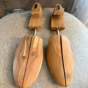 May include: A pair of light brown wooden shoe trees featuring a split-toe design, a central metal spring mechanism, and a rounded heel piece. The natural wood grain is visible across the surface of both shoe trees, which are shaped to fit inside footwear to maintain their form.