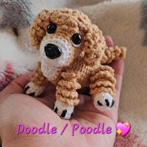 May include: A handmade crochet dog figurine in shades of tan, white, and black. The dog has large black eyes and a white muzzle. The text "Doodle / Poodle" is visible in pink. The dog is resting on a soft, patterned surface.