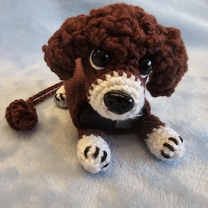 May include: A handmade crochet dog toy in brown and white. The dog has large black eyes, a white muzzle, and white paws with black details. The toy is lying down on a light blue surface.