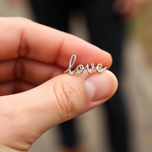May include: A silver ring spelling out the word "love" in a delicate, cursive font. The ring is held between fingers, with the word "love" prominently displayed. The ring's design is simple and elegant.