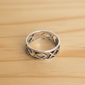 May include: A silver ring with a repeating fish symbol design. The ring is made of a shiny metal and has an openwork pattern. The ring is on a light brown wooden surface.