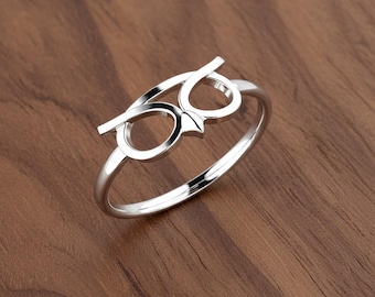 May include: A silver-colored ring featuring an owl design. The ring's design incorporates the shape of eyeglasses, with two circular lenses and a horizontal bar above. The owl's face is suggested by a small, pointed element between the lenses.