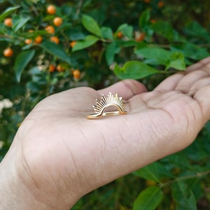 Puede incluir: Un anillo dorado con un diseño de sol. El anillo presenta una banda curva con líneas verticales que irradian hacia afuera, creando una apariencia de sol. El anillo se encuentra sobre un fondo de follaje verde y pequeñas bayas naranjas.