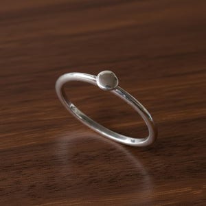 Puede incluir: Un anillo minimalista de plata con un pequeño acento circular. La banda del anillo es delgada y pulida, y el acento circular está centrado en la parte superior. El anillo se muestra sobre una superficie de madera oscura.