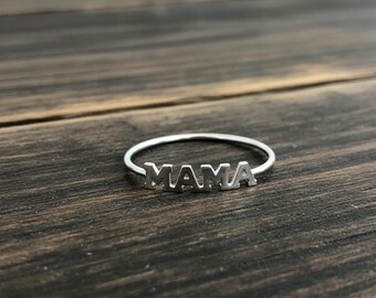 Könnte beinhalten: Ein silberner Ring mit dem Wort "MAMA" in Großbuchstaben. Der Ring ist dünn und liegt auf einer dunklen Holzoberfläche. Die Buchstaben sind ausgeschnitten, wodurch ein zartes Design entsteht. Der Ring ist ein Schmuckstück.